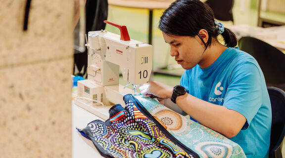 A person who is wearing a ligth blue shirt, and has dark hair tied back and is sewing at a table. A sewing machine is visible and a colourful piece of fabric.
