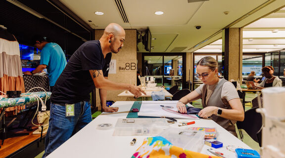 People sewing at The Edge during Threads 2024, someone is visible on the right hand side using a ruler to cut out a pattern, someone on the left hand side is standing over the table and pointing at the pattern to help.