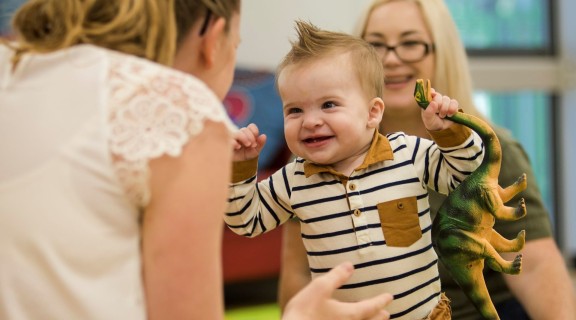 A baby and parent smile and play with a dinosaur toy.