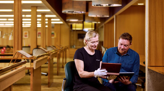 Man and woman sit closely, looking at an old case. The woman wears white gloves for handling.