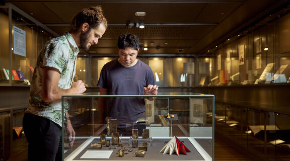 Two men view an artists’ book displayed in a glass case in a gallery space.