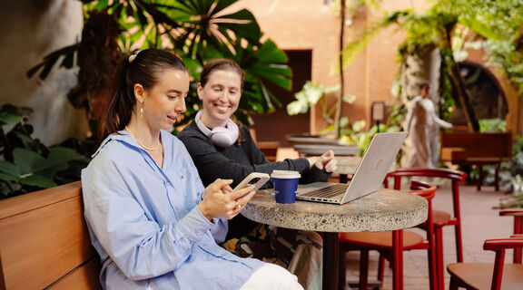 Two women at a cafe, one with a laptop and the other with a mobile phone.