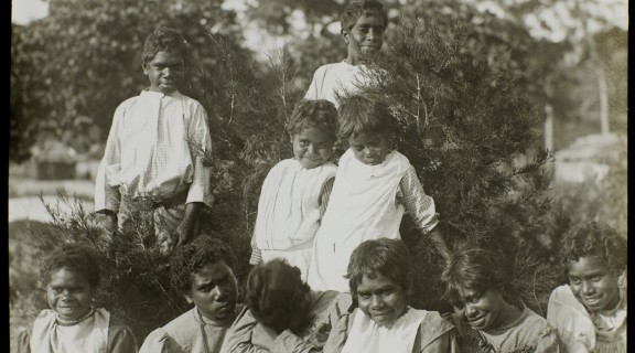 Group of First Nations children in front of shrubs, four children are standing with six children sitting on the ground in front.