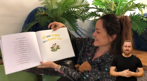 Library staff member reading a book with an Auslan interpreter also visible on screen