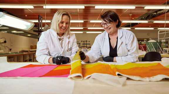 Two women wearing white lab coats and gloves carefully handling and preparing a fabric roll for preservation storage.