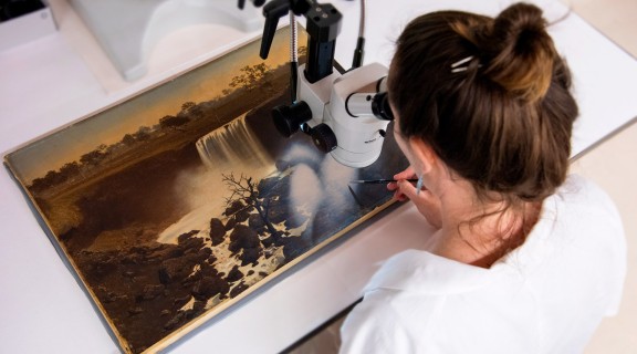 Over the shoulder shot of a State Library staff member looking at a painting through a microscope and painting small details with a paintbrush.
