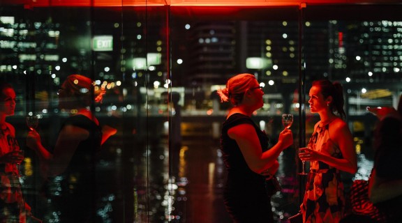 Two people conversing while holding drinks, illuminated by red light. The cityscape and river are visible in the background.