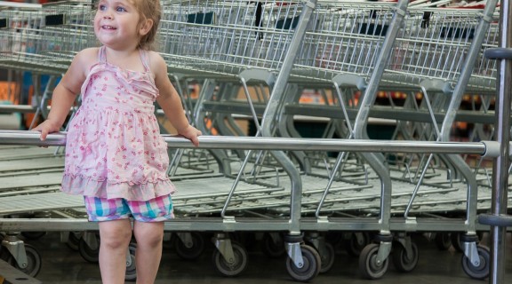A small child leans against a metal rack where shopping trolleys are lined up behind her. She is looking in the distance and looks happy