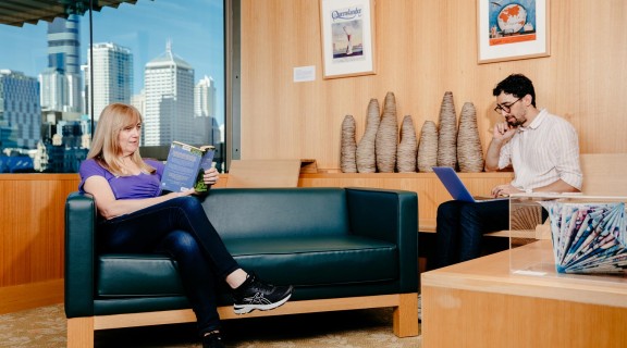 Two visitors in the John Oxley Library. One visitor reads a book on a couch while the other is using a laptop.