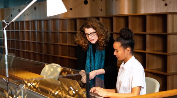 Woman gesturing to the screen of a laptop being used by a young person. A lamp is pointed towards them and behind them are wooden shelves.