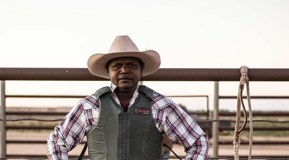An Aboriginal man dressed in rodeo protective gear stands in front of a fence with a rope hung next to him.