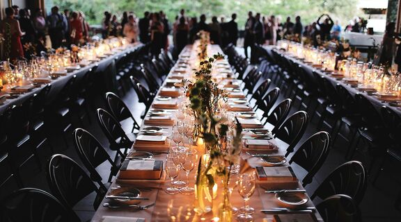 Long tables set with dinnerware, chairs, and flowers for a wedding on the Queensland Terrace, with a crowd of guests in the background.