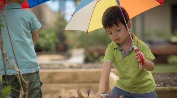 Two children are holding umbrellas. One is turned away from the camera. The other one is facing forward and looking down at a garden bed.