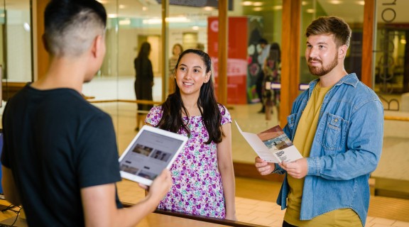 Two visitors talking to a State Library staff member at the reception desk. One visitor is holding a brochure.