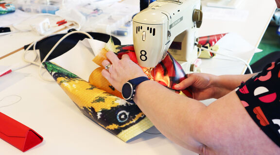 A women's arms are seen holding a brightly patterend piece of vinyl material, she is sitting in front of the showing machine and sewing the piece into a shape.