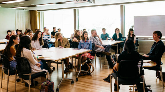 A group of people seated around square tables, attentively watching two speakers at the front of the room.