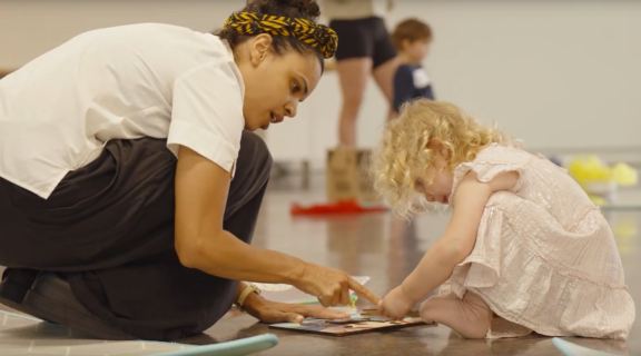 Women with patterned yellow headband points at a book with a blonde toddler.