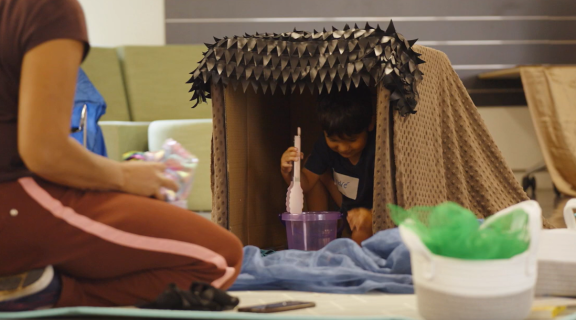 A small child plays with a bucket and tongs inside a cardboard box decorated with fabric and texture. 