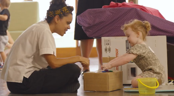 A lady in a white shirt and patterned yellow headband sits cross legged with a redheaded toddler playing with a box.