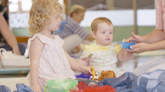 A toddler in a pink dress and an infant in a yellow shirt sit amongst fabric and toys. they are looking at a toy shark.