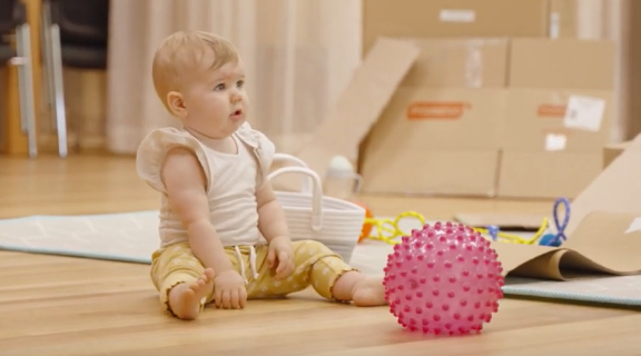 A baby in a white shirt and yellow trousers looks curiously at something outside of frame. A pink plastic ball with bumps on it sits in front of them.