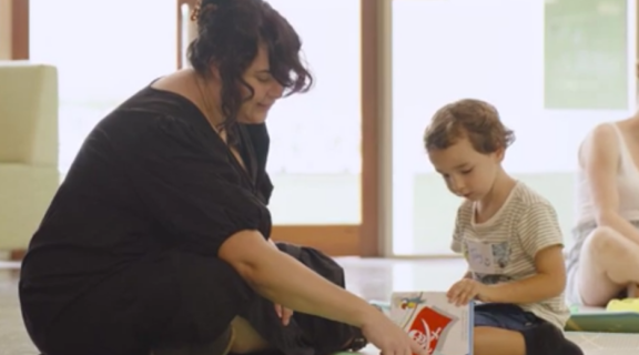 a woman with dark hair points to a page in a book a toddler is holding. 
