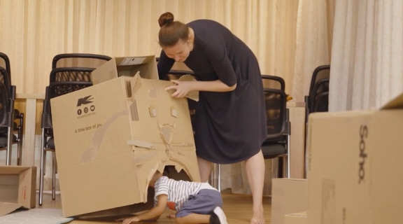 A child in a striped shirt peaks out from underneath a cardboard box decoratd to look like a cat's face. A woman in a dark dress holds the box up for them.