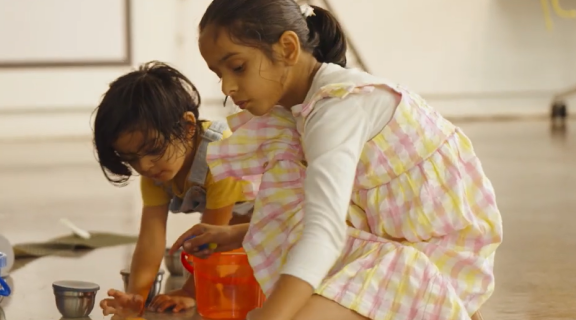 Two little girls, one in a yellow and pink dress, the other in yellow, play with counters and a bucket on the floor.