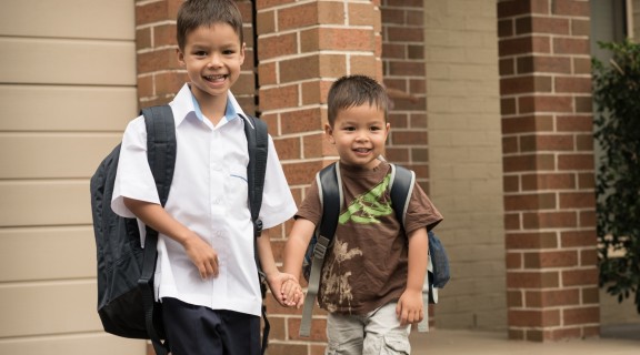 Two boys are holding hands. The older boy is dressed in a school uniform and carrying a large backpack. They are both smiling.