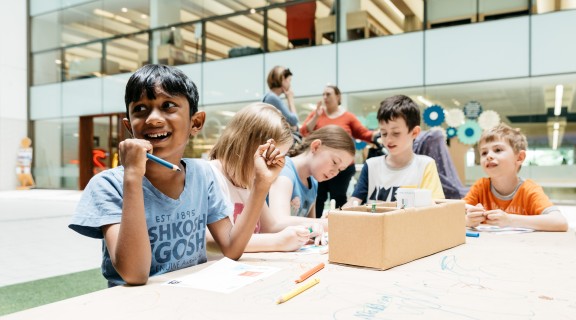 Five children drawing on tables at State Library of Queensland with two adults in the background.