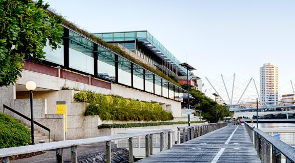 View of the exterior of the State Library building from the Brisbane River with Kurilpa Bridge and buildings in the background. 