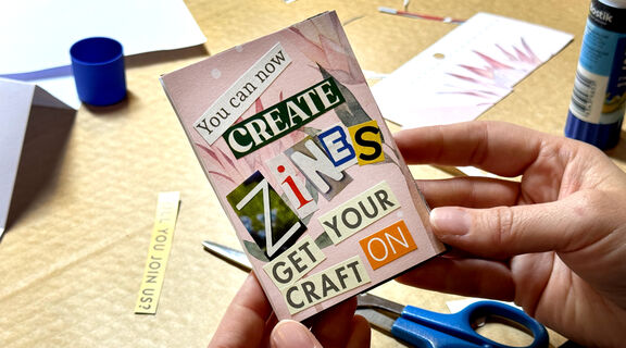 A person holds a handmade mini zine that says 'you can now create Zines' with cut out writing on a pink background