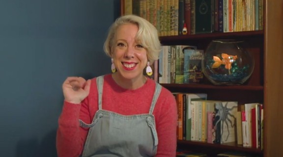 Woman in overalls and pink jumper waving