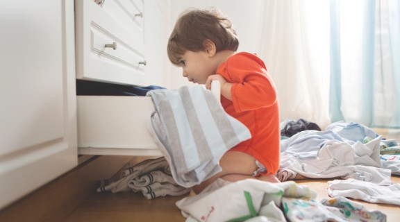 A toddler in an orange jumpsuit is looking into an open clothes drawer. There are clothes strewn across the floor.