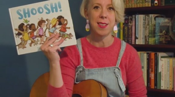 Lady in pink jumper with guitar, holding up Shoosh book