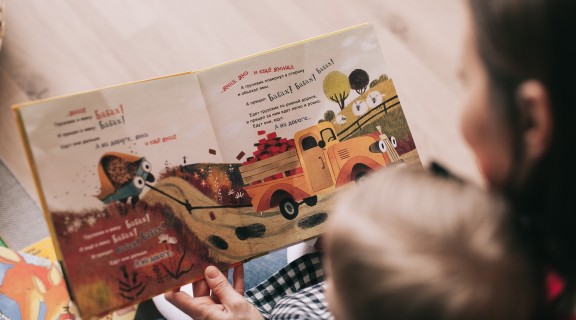 A woman is reading a picture book with a child. The book's text is in a Cyrillic language.