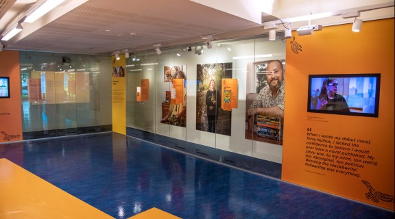 Inside the Sovereign Stories showcase at State Library. Large portraits of featured authors and publishers are on the wall behind glass and two screens are playing recorded interviews.