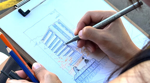 Persons hand holds a pen that is mid sketch of a city building in blue ink.