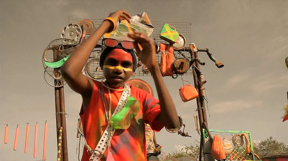 A young Indigenous person poses with arms up in neon face paint in front of a decorated arch. The aesthetic is 'neon junk punk'.