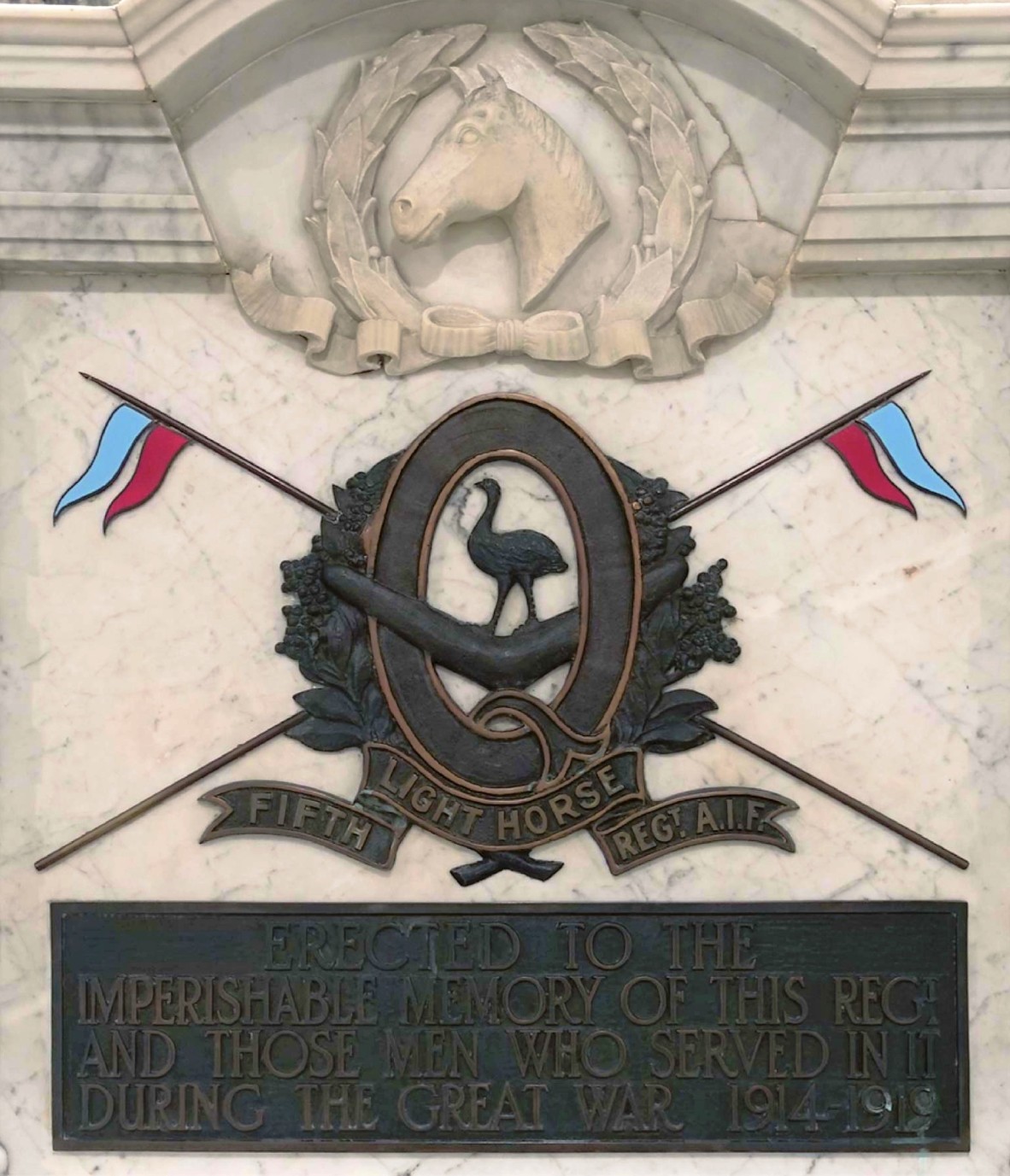 Dark brown plaque of letter Q surrounded by wattle wreath with emu in centre standing on boomerang blue and red flags crossing in the background Words below and white horse head in wreath above