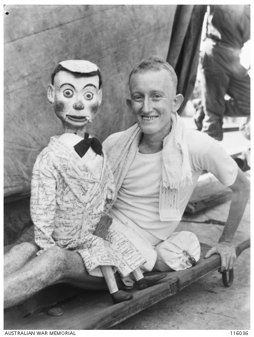 Gunner Thomas Hussey and “Joey” ventriloquist doll, photographed sitting on a stretcher after release from Changi prison, 1945.