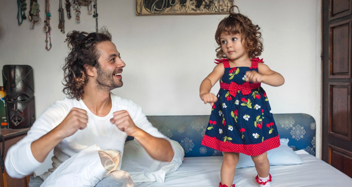 Girl dancing on bed while dad kneels beside, also dancing
