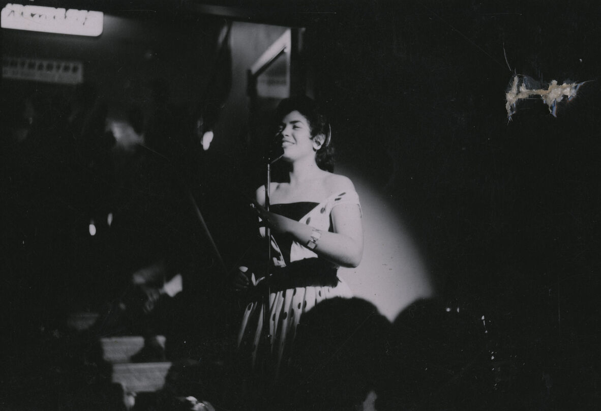 Dark-haired woman singing in front of microphone in dim setting with stage light behind her