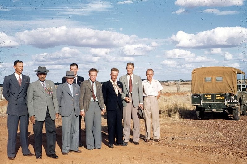 Group of veterans wearing suits and medals standing outside in Boulia near a Landrover in 1954 