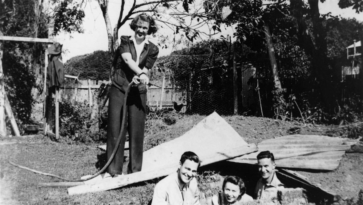 black and white photograph of Doreen Smythe standing above an air-raid shelter filled with friends in her backyard in Manly, Queensland.  