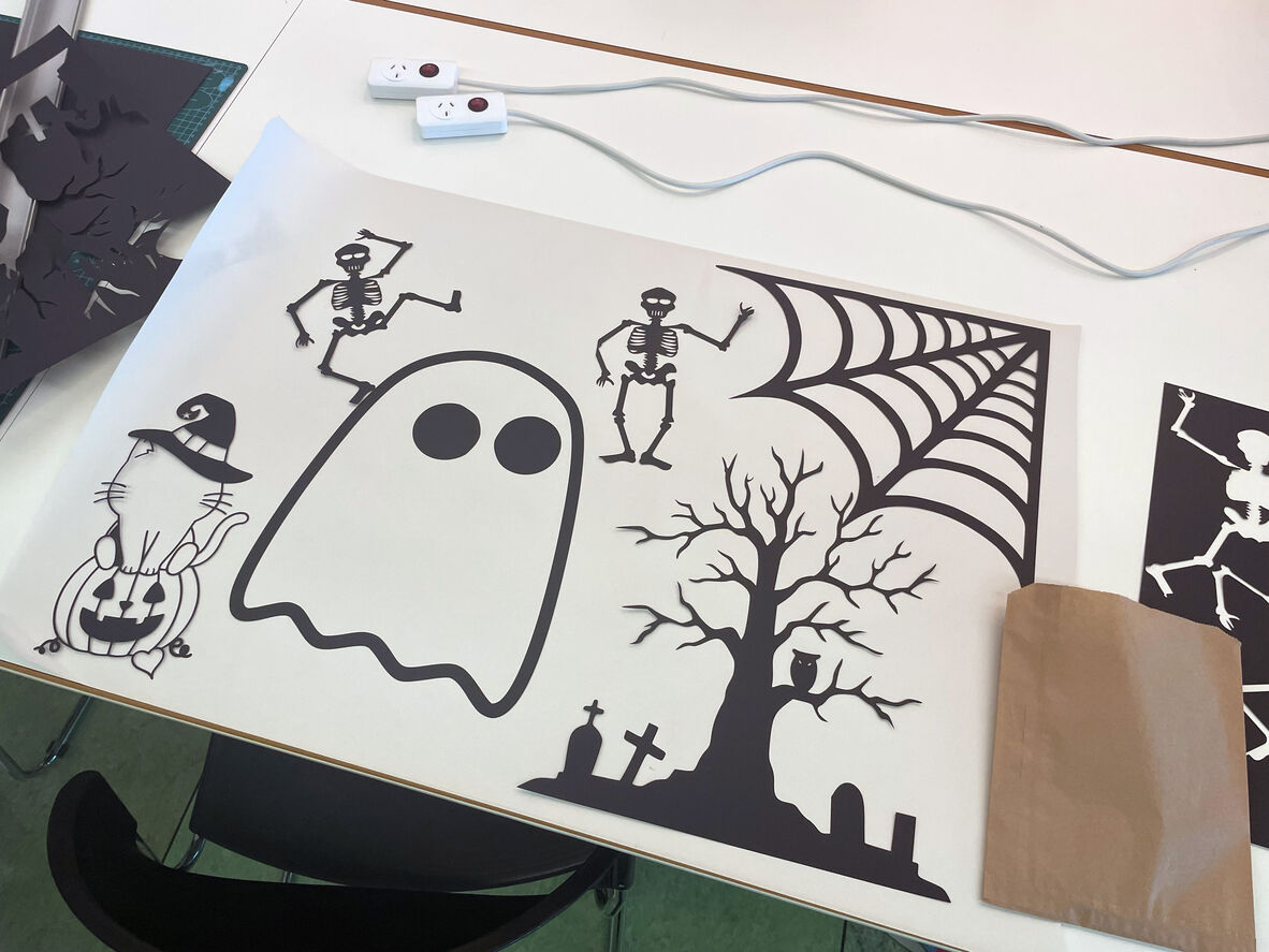 a bunch of halloween themed black laser cut outs on a white background. Two skeletons, a cobweb, a cat onto of a jack O lantern with a witch's hat, a ghost with big round eyes and a spooky looking tree in a graveyard.