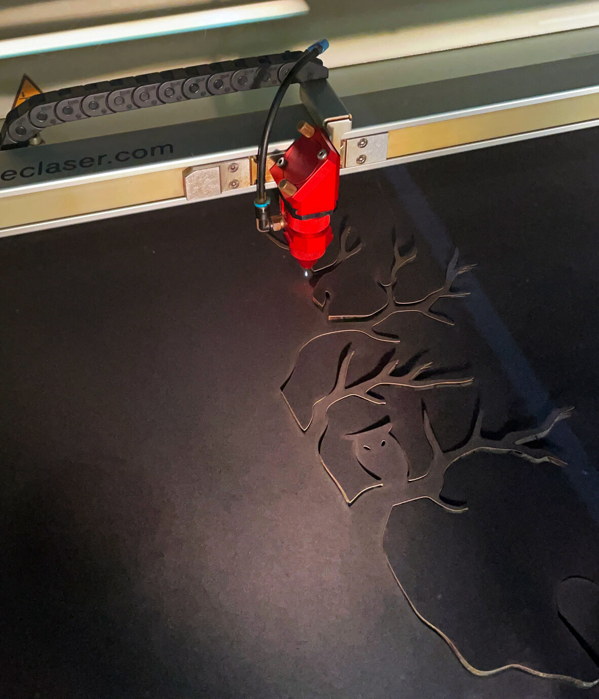 A laser cutter is carving a spooky tree design with branches and an owl shape into a dark sheet of material.