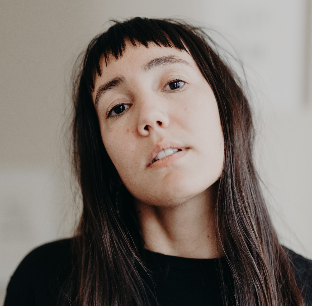 A person with long brown hair looking at the camera with head tilted back. They are wearing a black shirt against a white background.