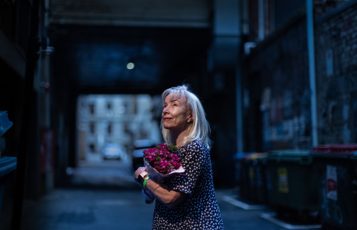 A photo of a women standing in an alleyway holding a bouquet of purple flowers