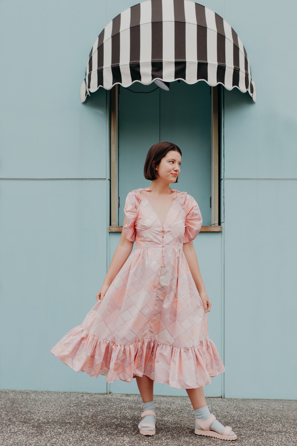 Kay Kerr stands in front of a blue wall. She wears a long pink dress and pink sandals. Photo by Jess Kearney.